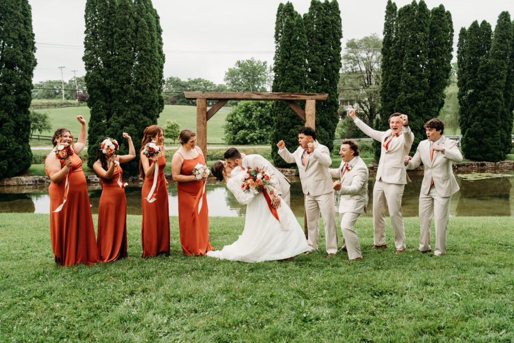 A wedding party of bridesmaids in red dresses and groomsmen in cream suits poses with the bride and groom in a manicured garden setting.